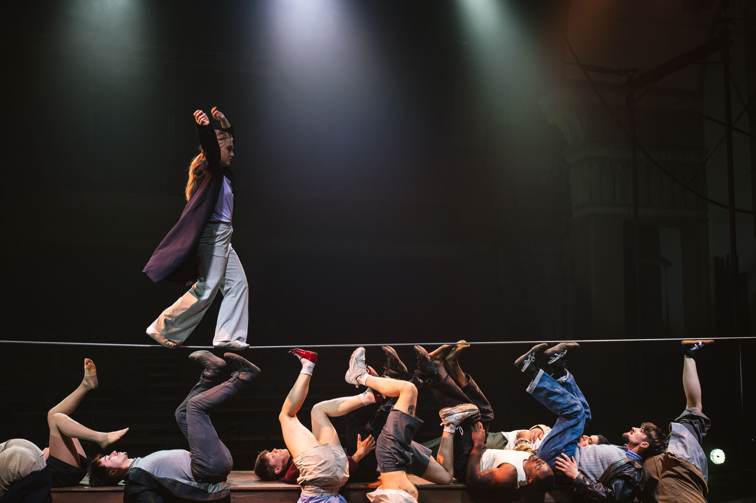 Artiste de cirque marchant sur un fil tendu au-dessus de plusieurs interprètes allongé·es sur scène, sous des éclairages dramatiques.